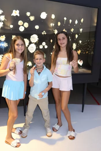 Three children standing in front of a lit display of varied shells at a museum, each holding a shell and smiling at the camera.